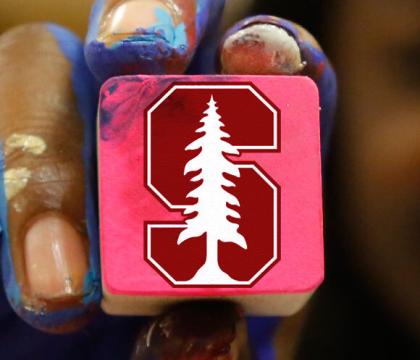 A paint covered hand holds a square block with the Stanford "S" insignia.